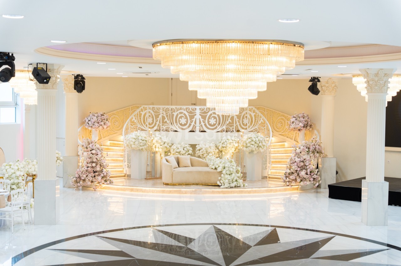 Salle de réception élégante avec escalier orné, canapé beige et décor floral abondant sous un grand lustre.