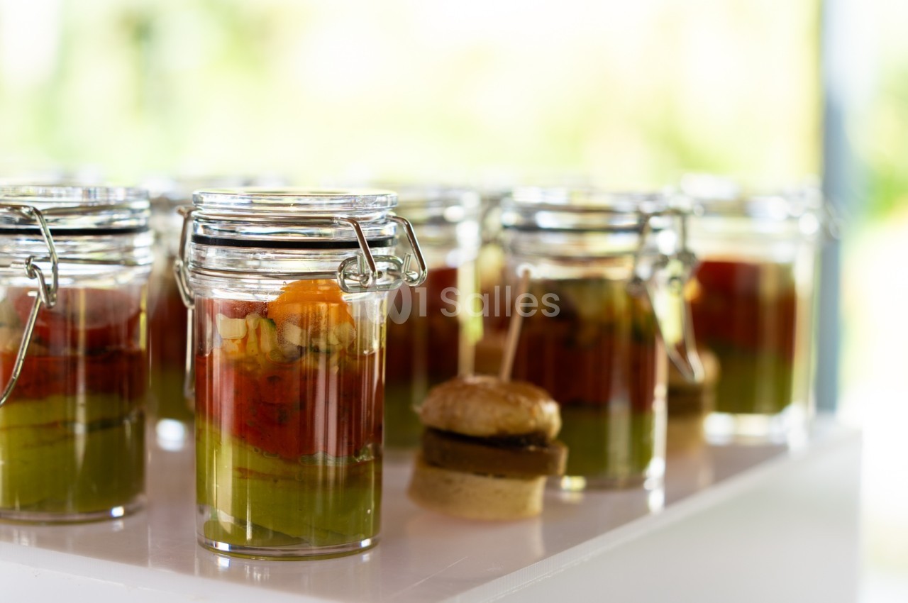 Verrines colorées avec couches de purées de légumes et mini-burger en arrière-plan sur une table blanche.