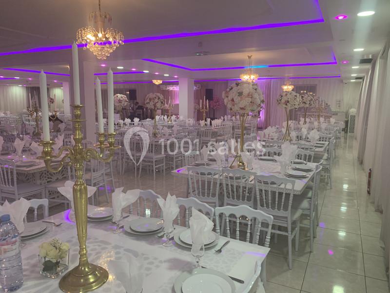 Salle de réception élégante avec tables dressées, chandeliers dorés, nappes blanches et éclairage violet.