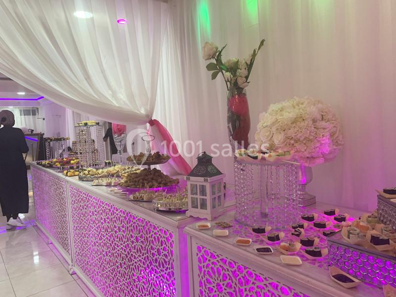 Buffet décoratif avec desserts variés, lanternes et fleurs blanches dans une salle éclairée par des lumières colorées.