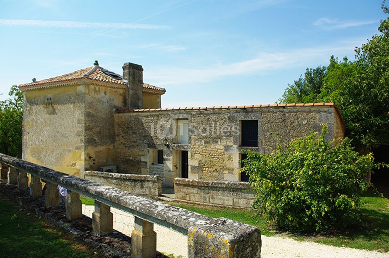 Bâtisse en pierre avec toit en tuiles, entourée de verdure et d'un muret, sous un ciel bleu dégagé.