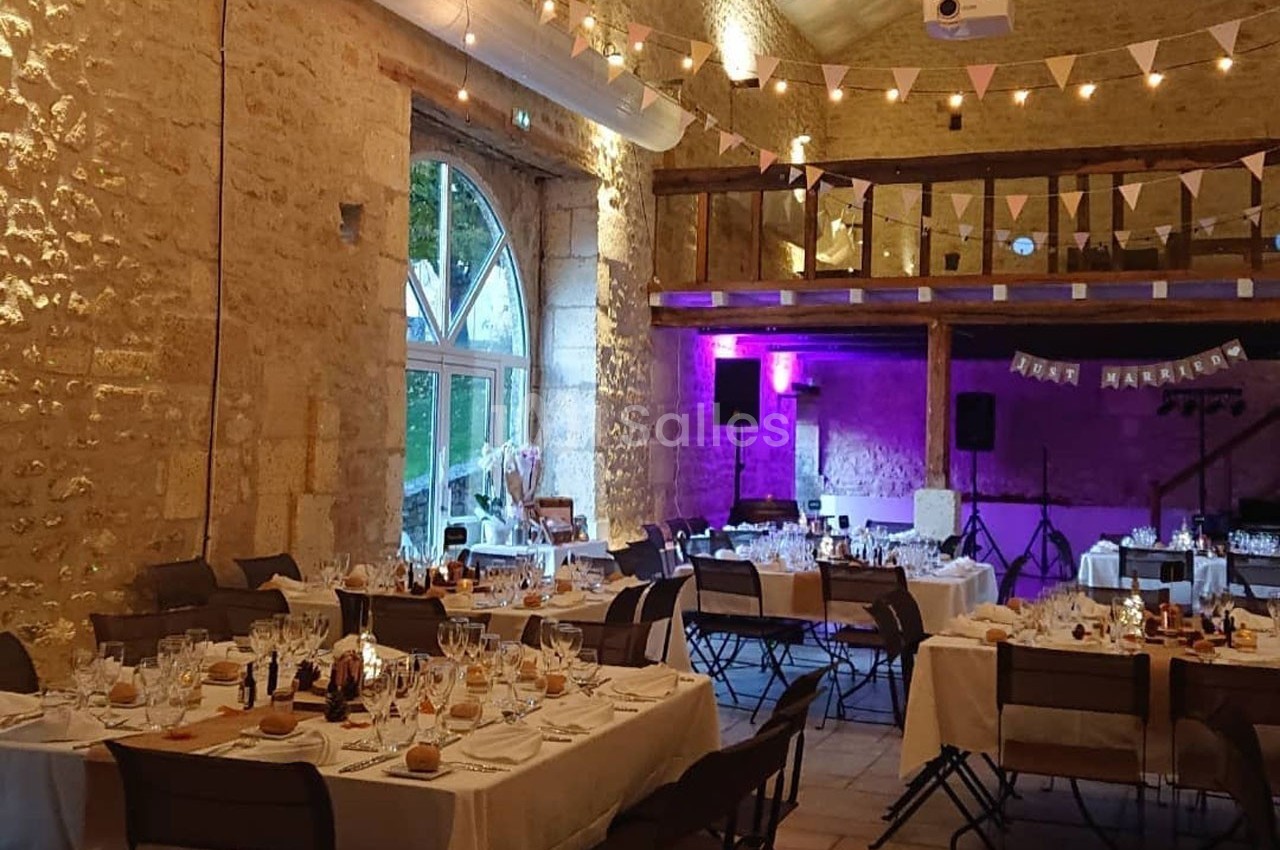 Salle de réception décorée pour un mariage, avec tables dressées, guirlandes lumineuses et murs en pierre apparente.