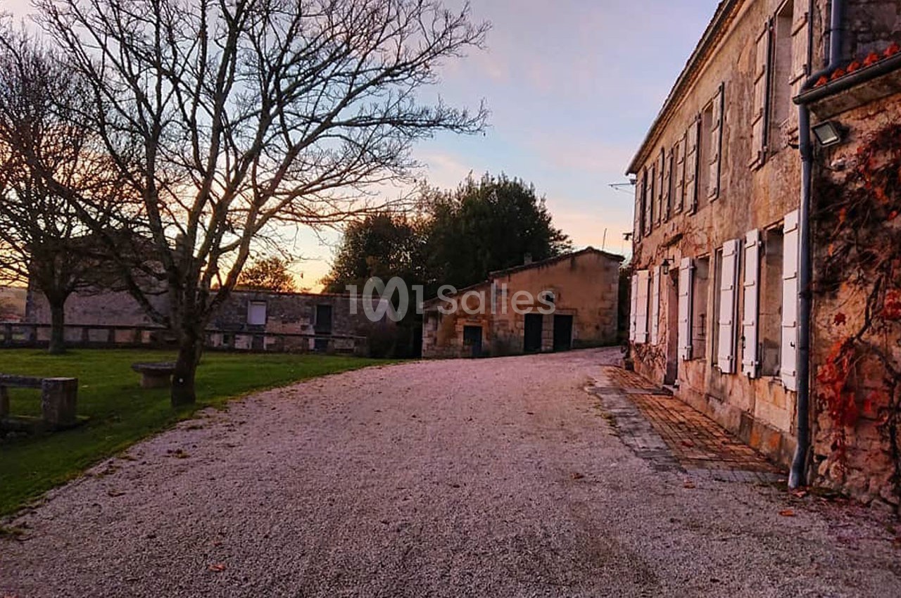 Cour pavée bordée de bâtiments en pierre, avec un arbre dénudé et un ciel aux teintes de coucher de soleil.