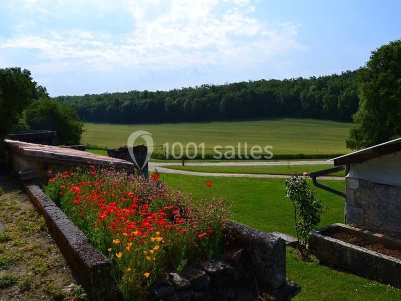 Champ verdoyant bordé de forêt, avec des coquelicots en premier plan et des bâtiments en pierre sur les côtés.