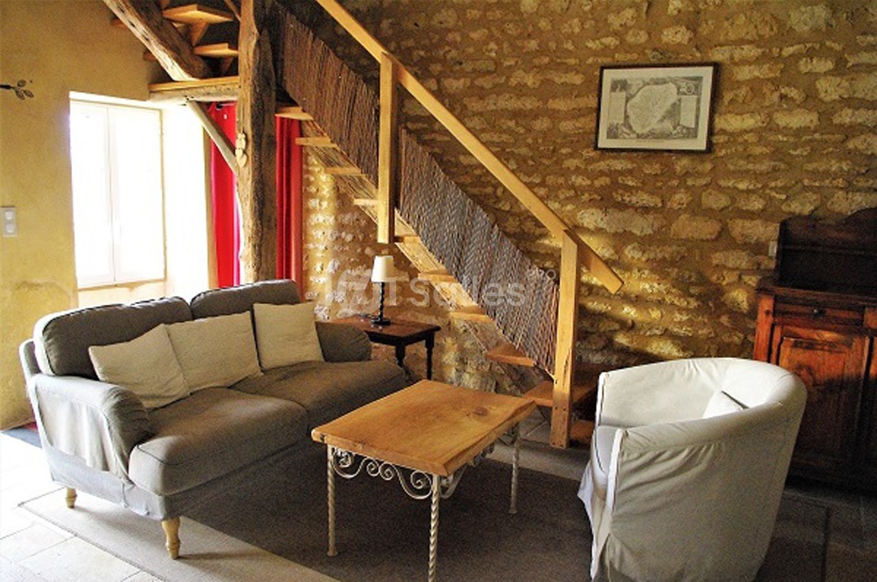 Salon rustique avec murs en pierre, canapé, fauteuil, table basse en bois et escalier en bois menant à l'étage.