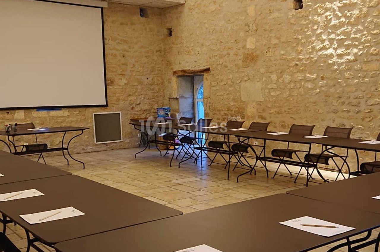 Salle de réunion en pierre avec tables disposées en U, chaises, écran de projection et lumière naturelle.