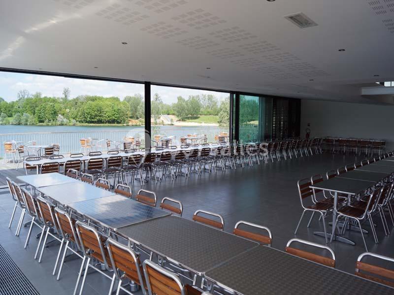 Salle spacieuse avec de grandes baies vitrées donnant sur un paysage verdoyant et un plan d'eau, tables et chaises alignées.