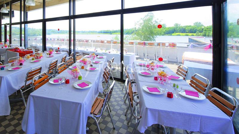 Salle de réception lumineuse avec tables dressées, nappes blanches, serviettes roses et vue sur un paysage extérieur.