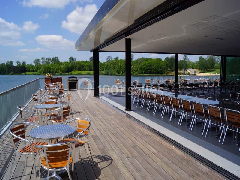 Terrasse en bois avec tables et chaises en métal, donnant sur un lac entouré de végétation sous un ciel dégagé.