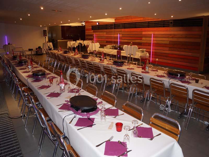 Salle de réception avec de longues tables dressées, nappes blanches, serviettes violettes et chaises en bois.
