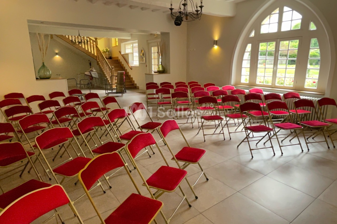 Salle lumineuse avec des chaises rouges alignées, un escalier en bois et de grandes fenêtres donnant sur un jardin.