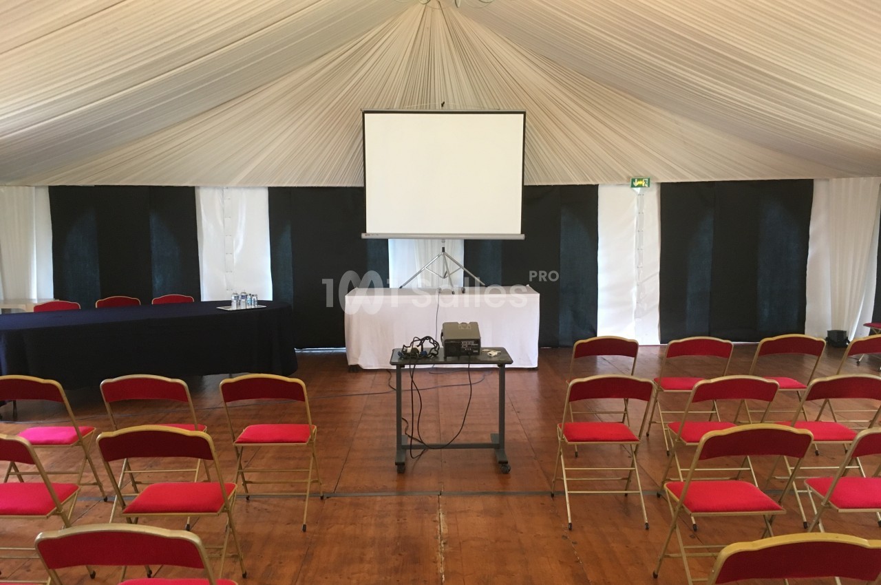 Salle de conférence avec chaises rouges alignées, écran de projection et matériel audiovisuel sur un chariot.