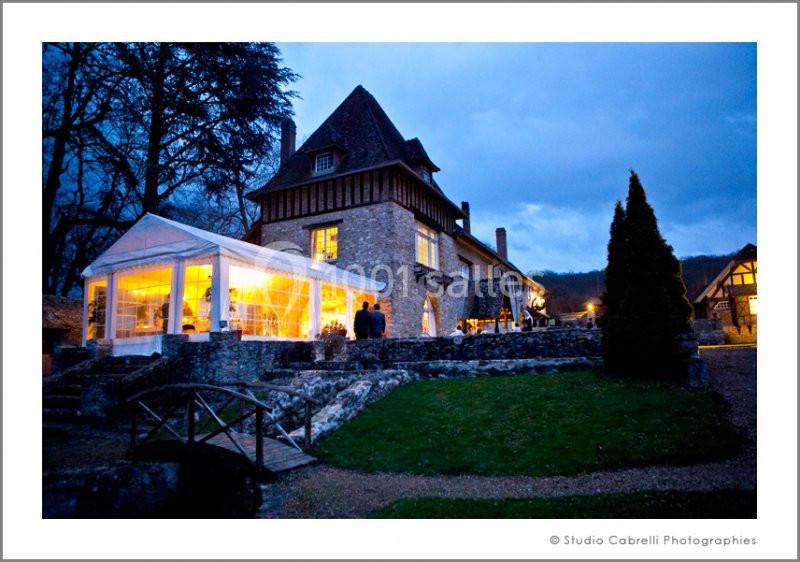 Grande maison en pierre avec une verrière éclairée, entourée d'arbres et d'un jardin au crépuscule.