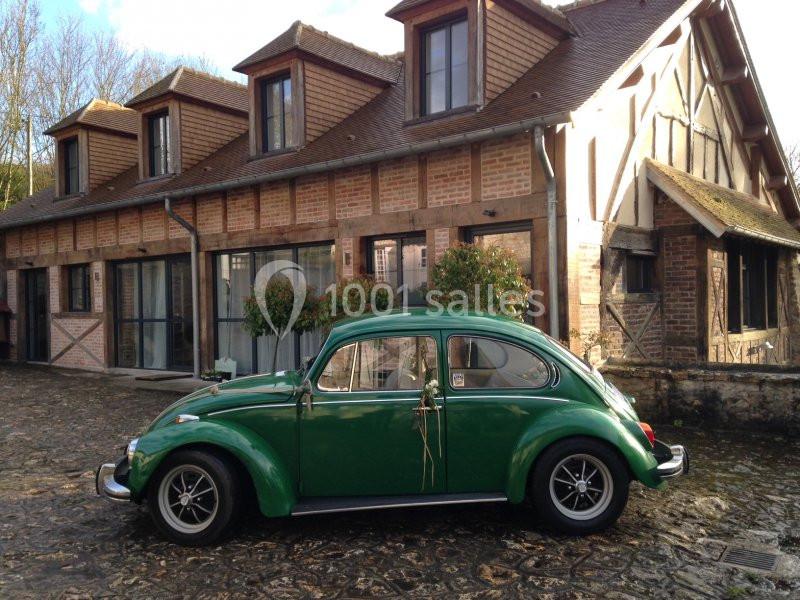 Voiture ancienne verte garée devant une maison en briques et bois à colombages, sur une cour pavée.