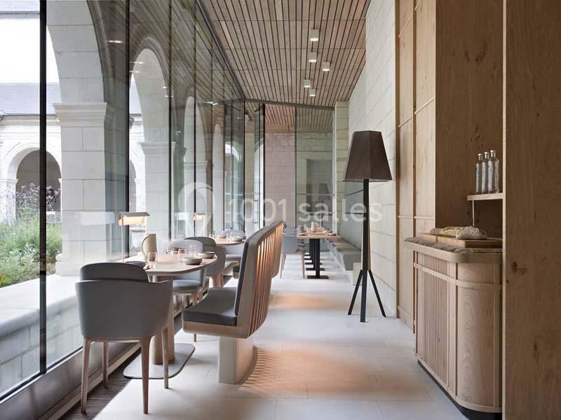Salle de restaurant lumineuse avec grandes baies vitrées, mobilier moderne en bois et vue sur un jardin extérieur.