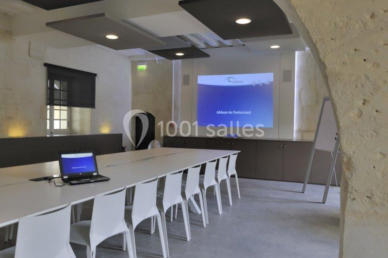 Salle de réunion lumineuse avec une grande table, des chaises blanches, un écran de projection et un ordinateur portable.