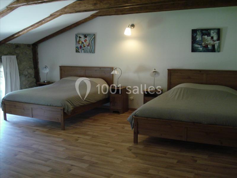 Deux lits doubles avec des draps gris dans une chambre spacieuse au sol en parquet, éclairée par une lampe murale.