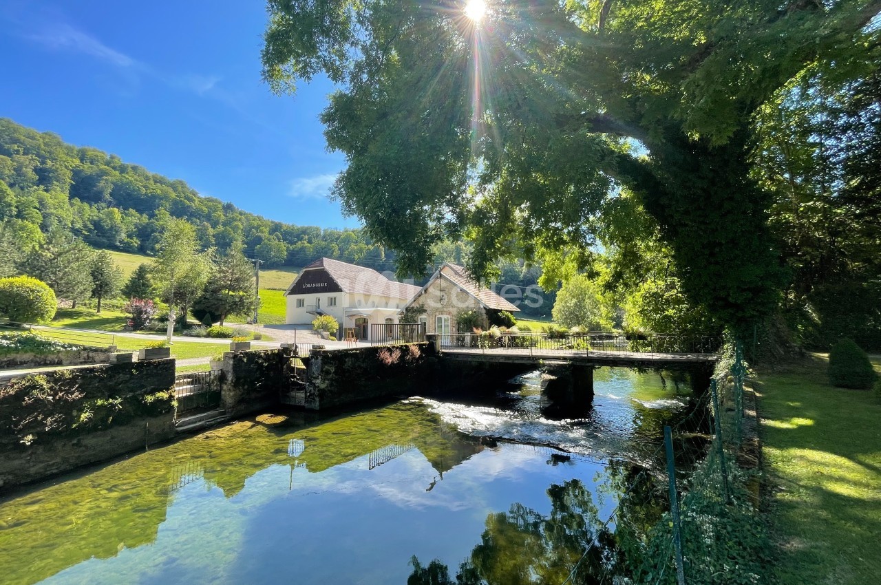 Maison au bord d'une rivière avec un pont, entourée de verdure et éclairée par un soleil traversant les arbres.