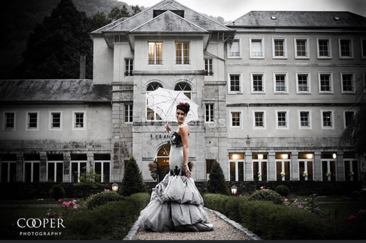 Femme en robe de soirée tenant un parapluie devant un bâtiment historique entouré de jardins.