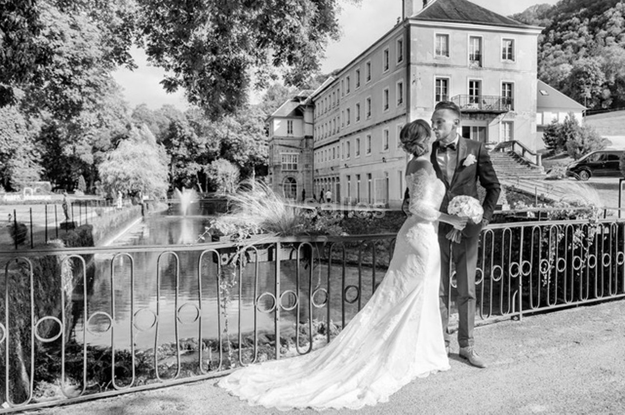 Un couple en tenue de mariage pose près d'un pont avec un bâtiment historique et un étang en arrière-plan.