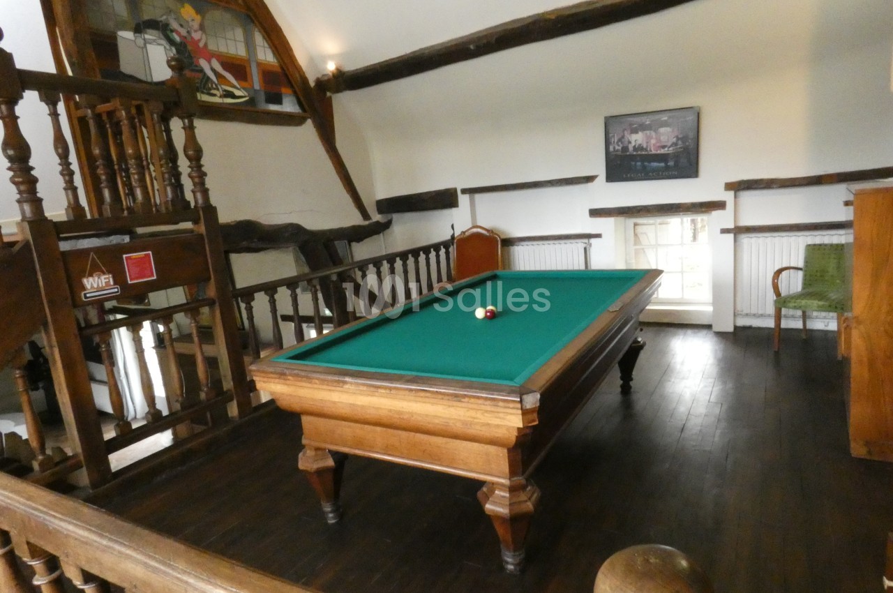 Table de billard en bois avec tapis vert dans une pièce à poutres apparentes et parquet sombre.