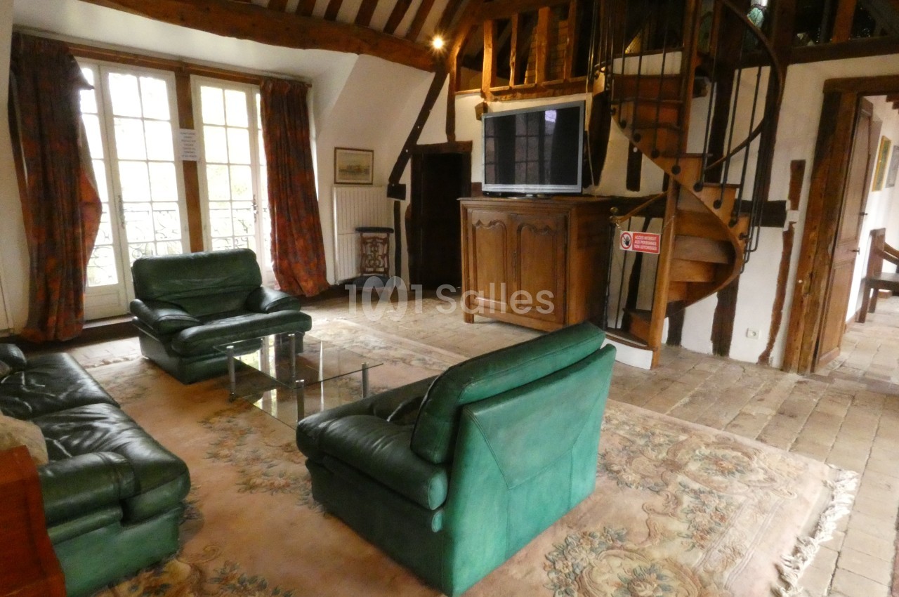 Salon avec poutres apparentes, meubles en bois, canapé vert, tapis beige et escalier en colimaçon en arrière-plan.