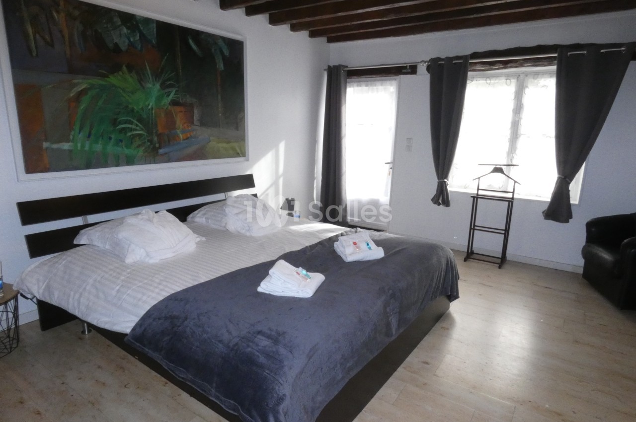 Chambre lumineuse avec un grand lit double, draps blancs, couverture grise, poutres apparentes et grandes fenêtres.