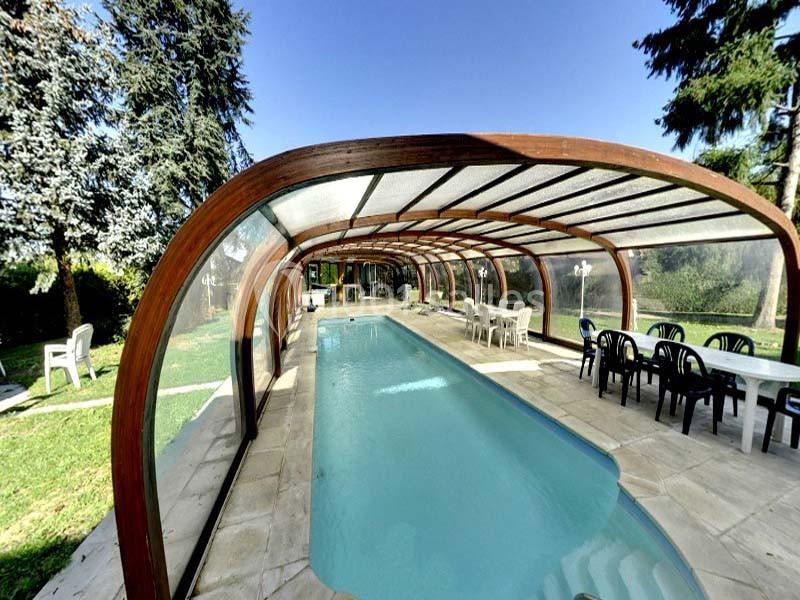 Piscine couverte par une structure en bois et verre, entourée de chaises et tables dans un jardin verdoyant.