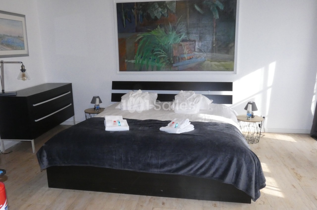 Chambre lumineuse avec un lit double, draps pliés, tableau mural, commode noire et sol en bois clair.