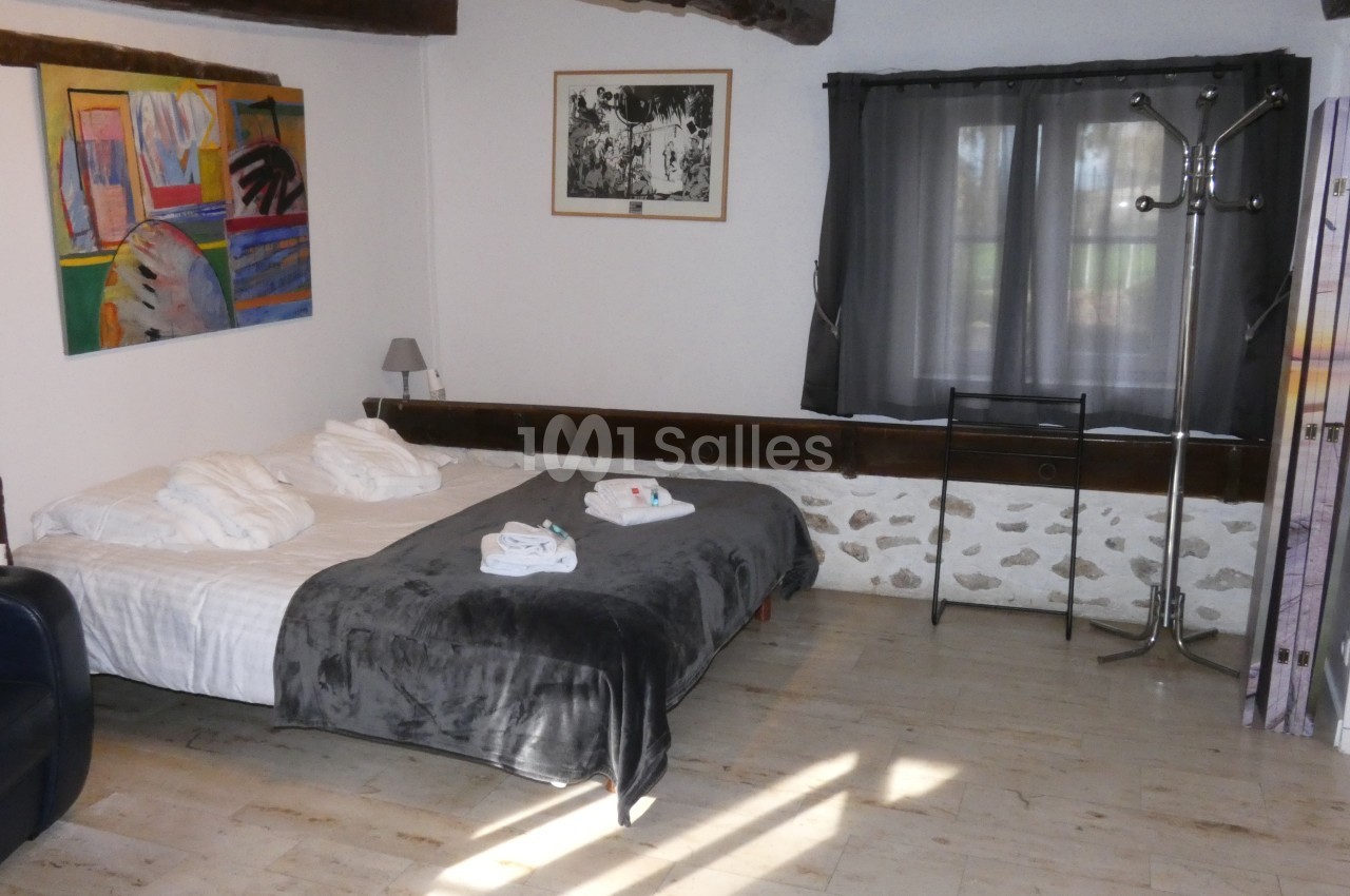 Chambre avec lit double, murs décorés de tableaux, poutres apparentes, sol en bois clair et fenêtre avec rideaux sombres.