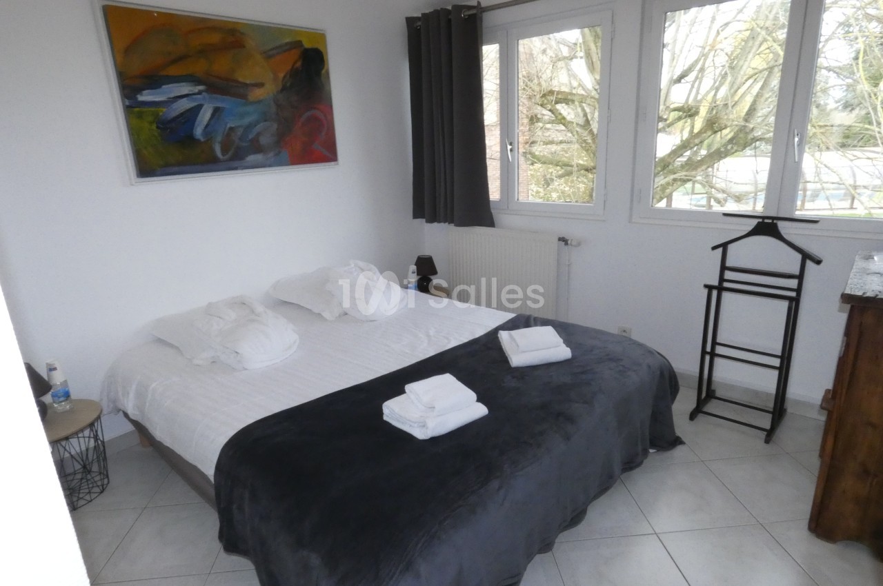 Chambre lumineuse avec un lit double, draps blancs, plaid gris, tableau coloré au mur et vue sur des arbres par la fenêtre.