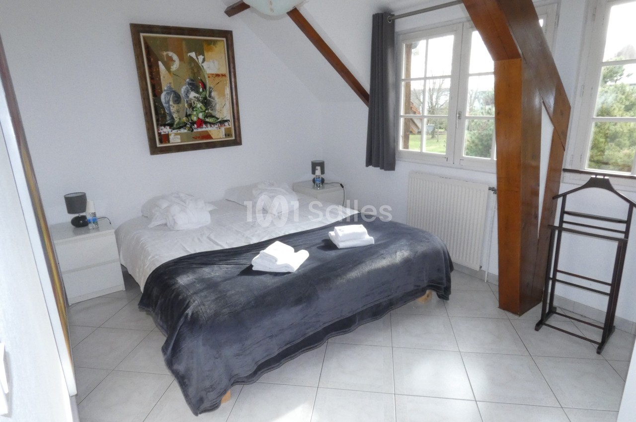 Chambre lumineuse avec lit double, couverture sombre, peignoirs pliés, grande fenêtre et poutres apparentes.