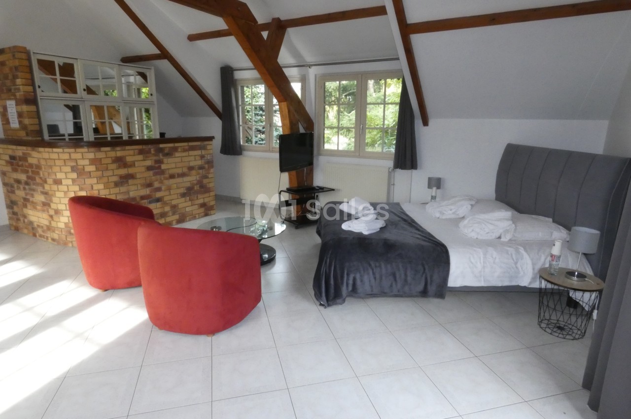 Chambre lumineuse avec lit double, fauteuils rouges, table basse, télévision et grande fenêtre donnant sur un jardin.