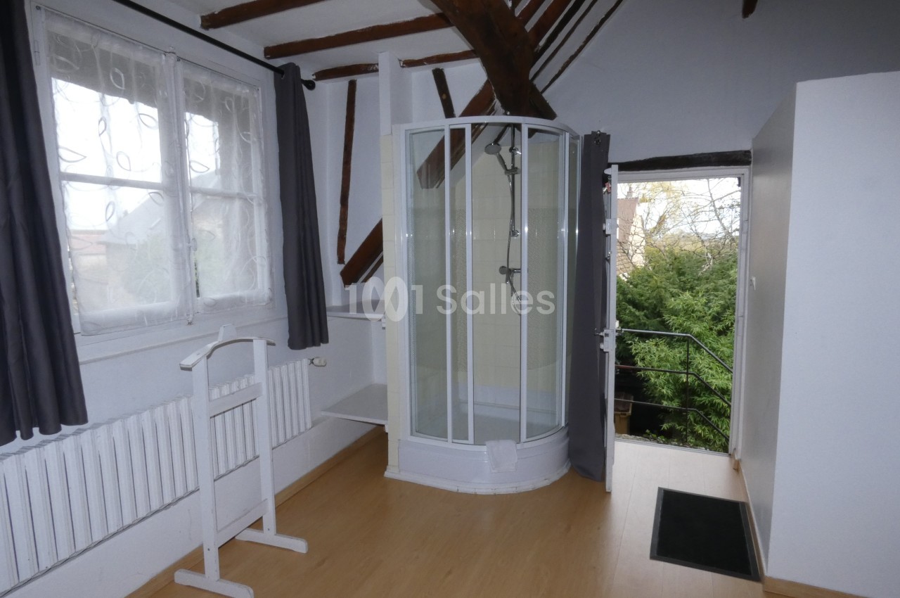 Chambre mansardée avec parquet, douche d'angle, fenêtre avec rideaux gris et vue sur un espace extérieur verdoyant.