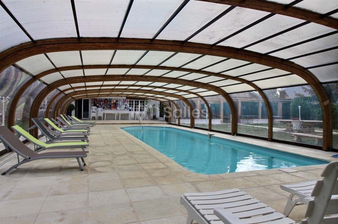 Piscine intérieure couverte avec structure en bois, entourée de chaises longues sur un sol carrelé.
