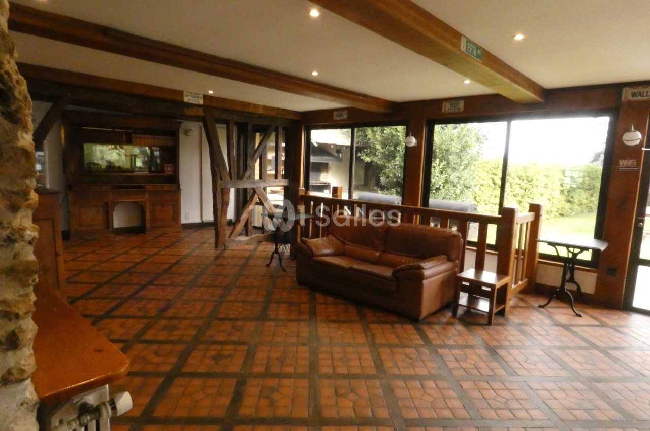 Salle lumineuse avec sol en carreaux, poutres apparentes, canapé en cuir et grandes baies vitrées donnant sur un jardin.