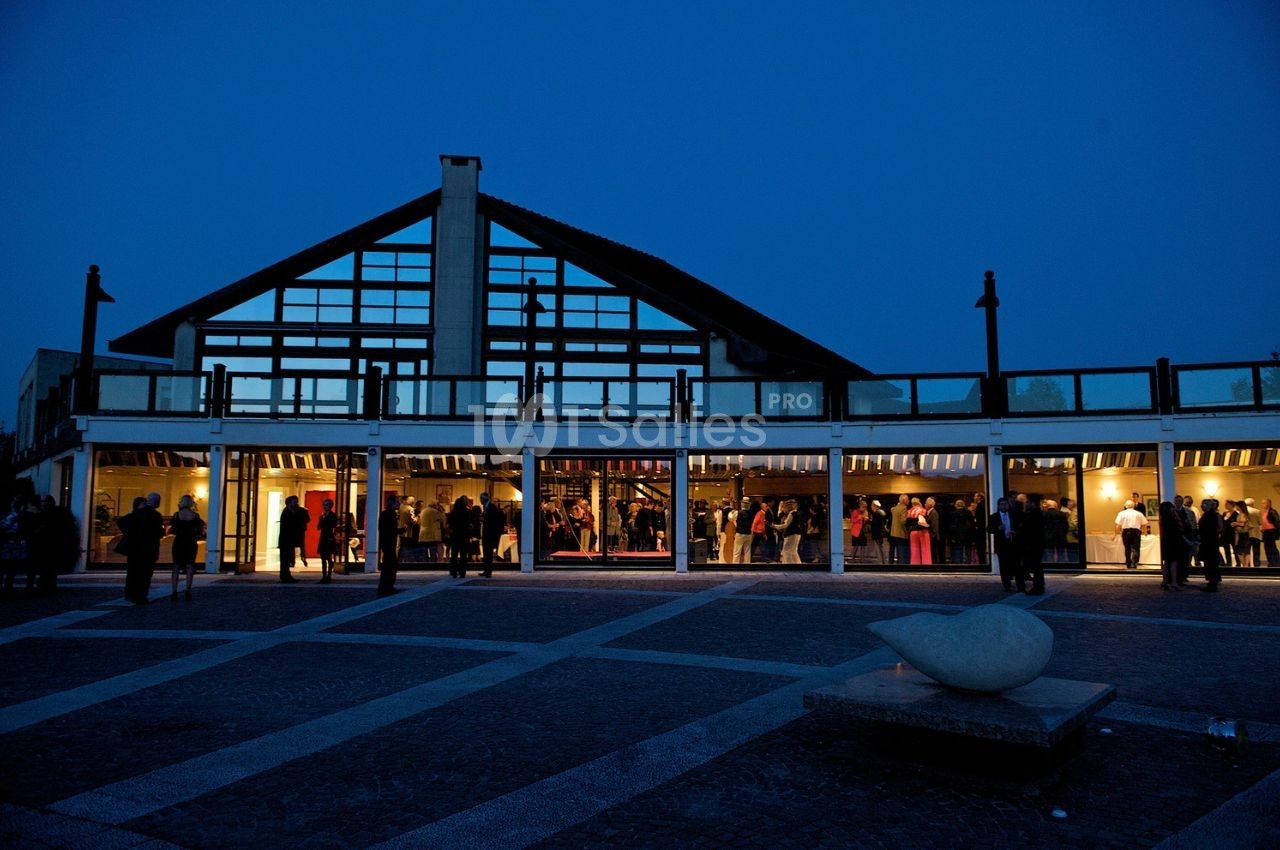 Façade vitrée d'un bâtiment illuminé en soirée, avec des personnes rassemblées à l'intérieur et à l'extérieur.
