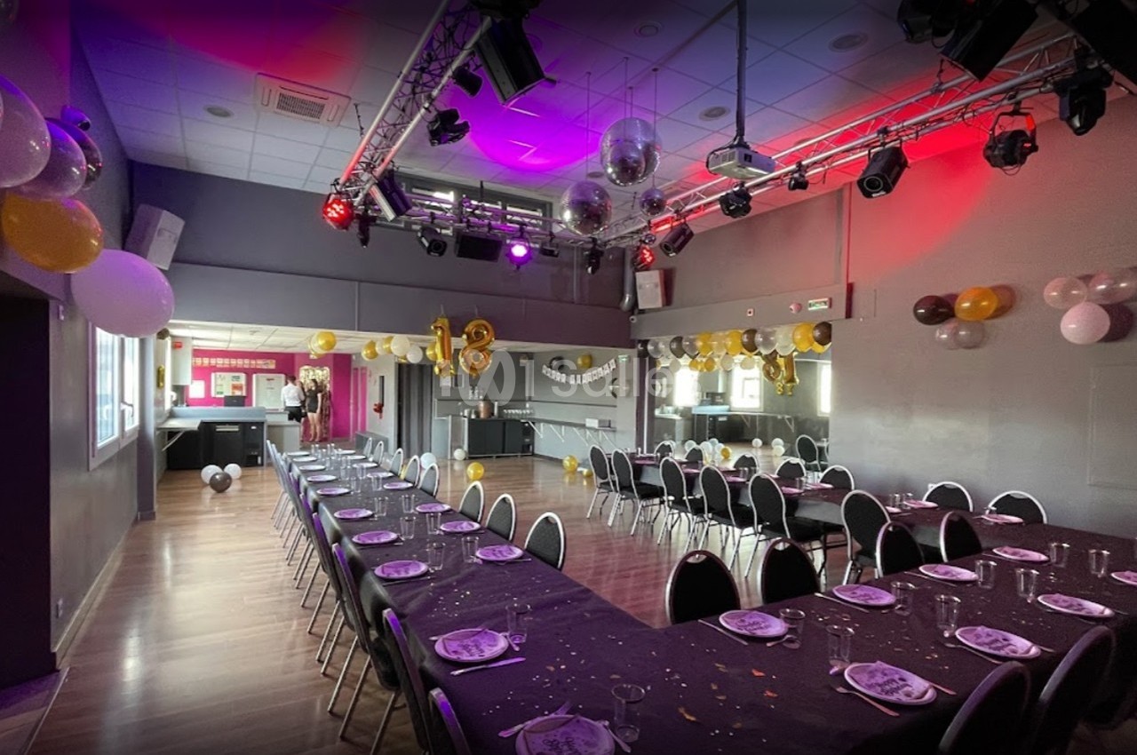 Salle décorée pour un événement, avec longues tables dressées, ballons colorés et éclairage festif.