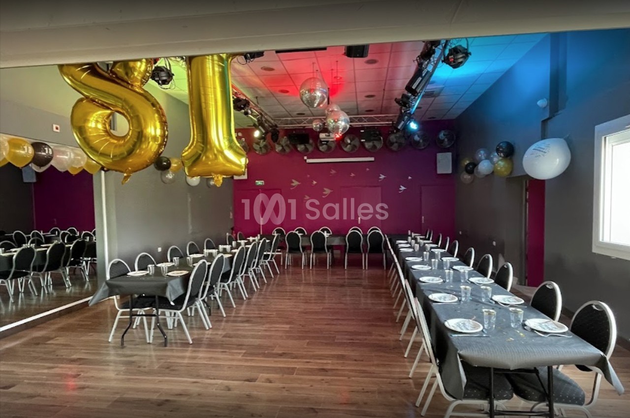 Salle décorée pour une fête avec ballons dorés, tables alignées avec vaisselle, et éclairage coloré au plafond.