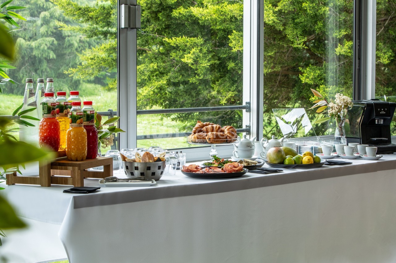 Buffet avec jus de fruits, viennoiseries, fruits frais, et machine à café, disposé près de grandes fenêtres donnant sur un…