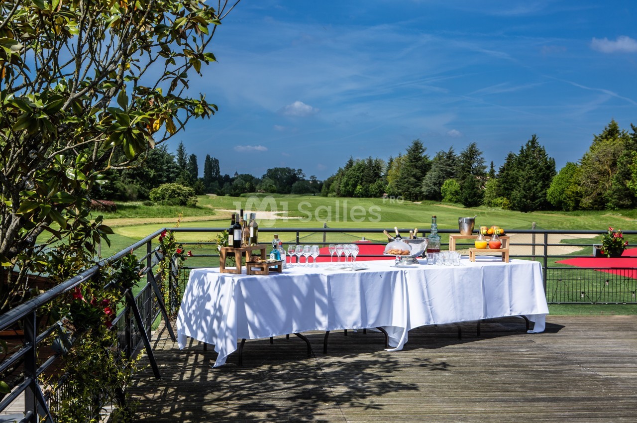 Table dressée en extérieur avec boissons et verres, sur une terrasse en bois surplombant un terrain de golf verdoyant.