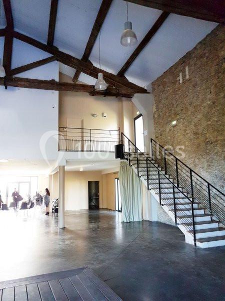Intérieur d'une salle avec murs en pierre, poutres apparentes, escalier métallique et mezzanine.