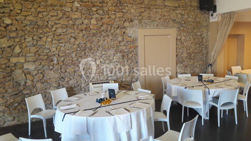 Salle avec murs en pierre, tables rondes dressées avec nappes blanches, chaises blanches et décorations simples.