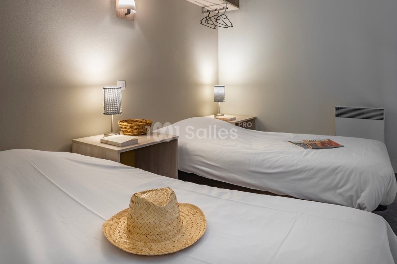Deux lits simples avec draps blancs, chapeaux en paille et lampes de chevet allumées dans une chambre sobre.