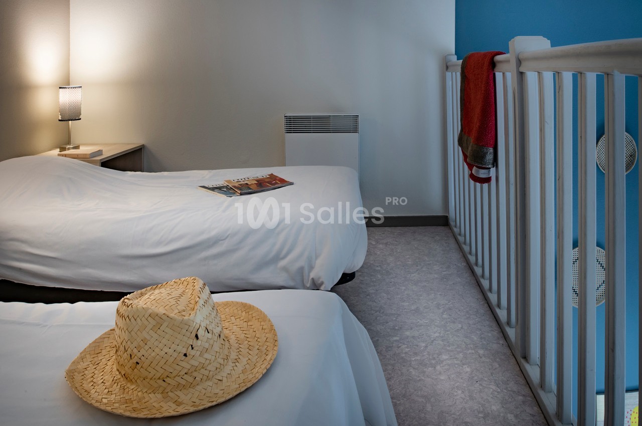 Deux lits simples avec draps blancs dans une chambre lumineuse, un chapeau de paille posé sur l'un d'eux.