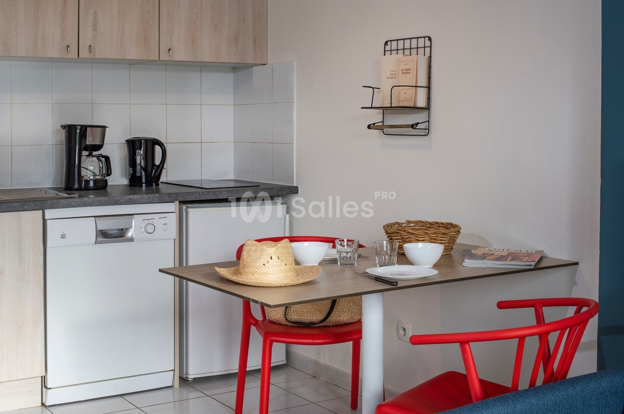 Cuisine avec table en bois, deux chaises rouges, vaisselle, cafetières, et étagère murale avec livres.