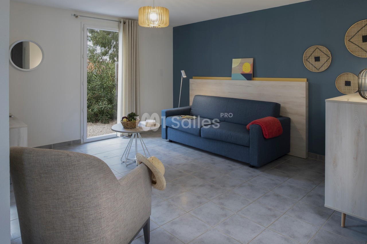 Salon lumineux avec canapé bleu, fauteuil beige, table basse ronde, mur bleu décoré et baie vitrée donnant sur un jardin.