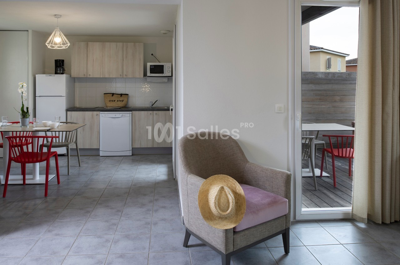 Salon avec fauteuil, chapeau posé, cuisine ouverte en arrière-plan et terrasse extérieure avec table et chaises rouges.