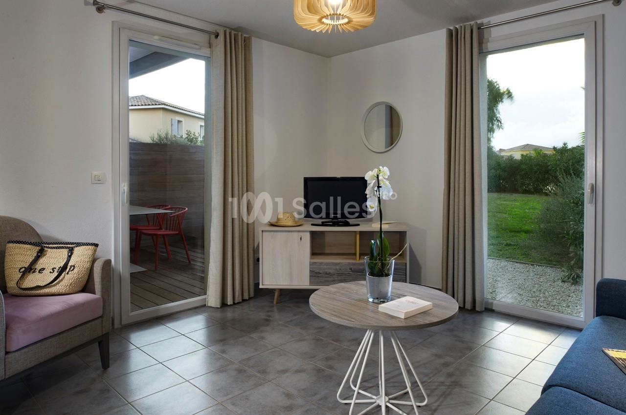 Salon lumineux avec table basse, orchidée, télévision sur meuble, deux portes-fenêtres donnant sur terrasse et jardin.