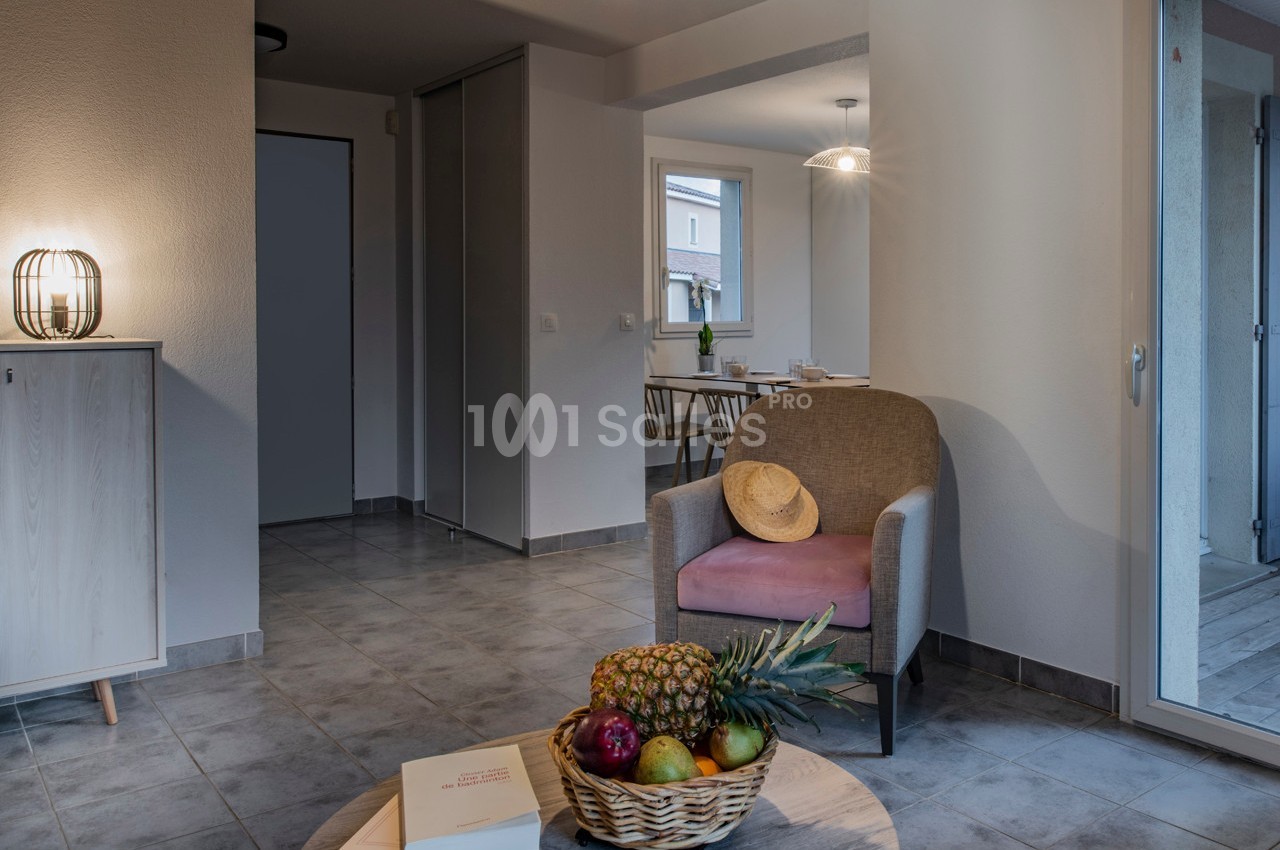 Salon lumineux avec fauteuil, panier de fruits sur une table basse et vue sur une salle à manger en arrière-plan.